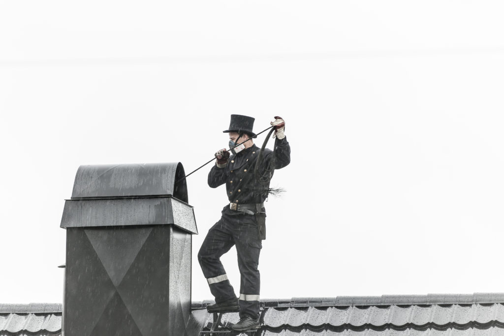 Bilde av en feier som står på et tak og feier en skorstein. Han er ikledd svart uniform med flosshatt.